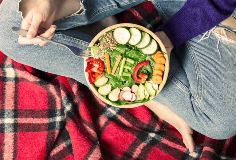 Healthy dinner or lunch. Woman in jeans sitting on red blanket with crossed legs and eating vegan in a bowl with vegetables, spinach, broccoli, seeds, fresh salad, carrots and avocado and word "veg" made from vegetables