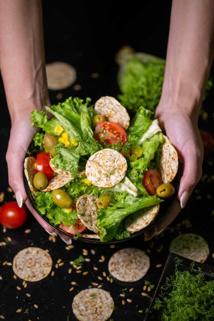 vertical closeup of a person holding a bowl of salad with crackers and vegetables under the lights